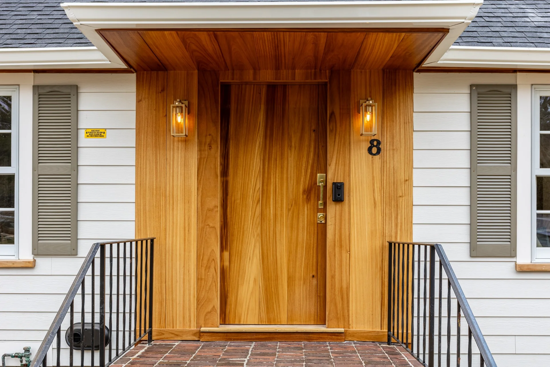 Custom wood front entry with warm paneling, exterior lighting, door trim, and a clean brick landing by Prospera Home Solutions.