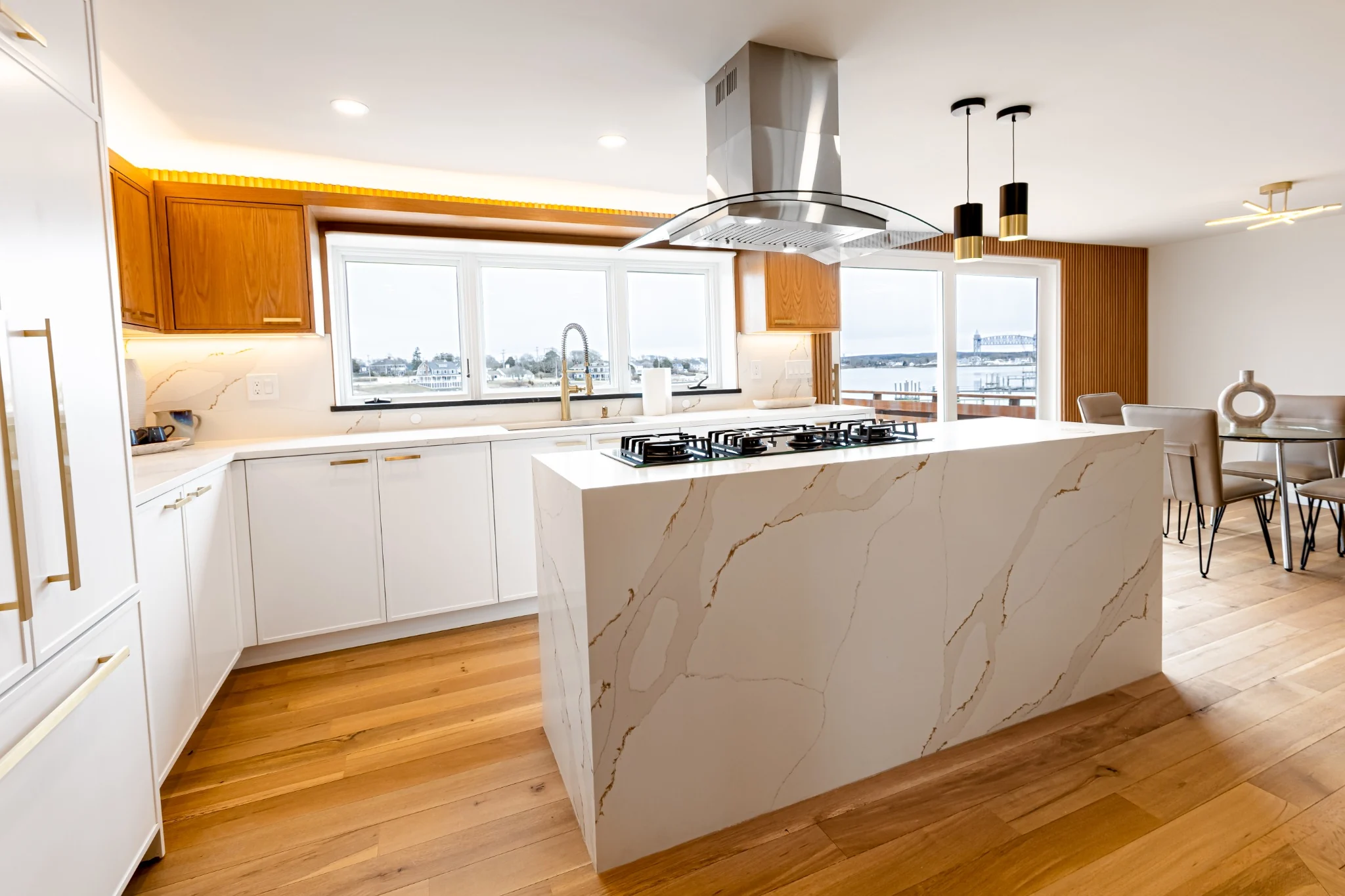 Modern waterfront kitchen renovation with a waterfall quartz island, mixed white and wood cabinetry, and large windows.